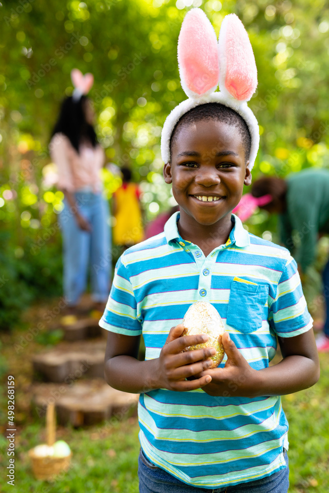Fototapeta premium Portrait of smiling african american boy in bunny ears holding easter egg while family in backyard
