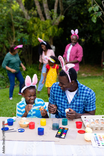 Happy african american boy and father painting easter eggs while family in background