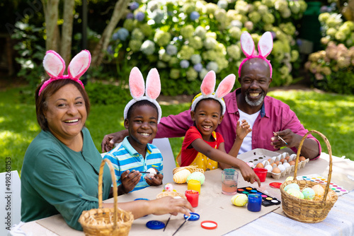Portrait of happy african american siblings and grandparents in bunny ears painting easter eggs