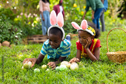 African american siblings in bunny ears arranging easter eggs on grass while family in background