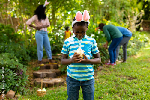 Happy african american boy in bunny ears looking at easter egg while family searching in backyard