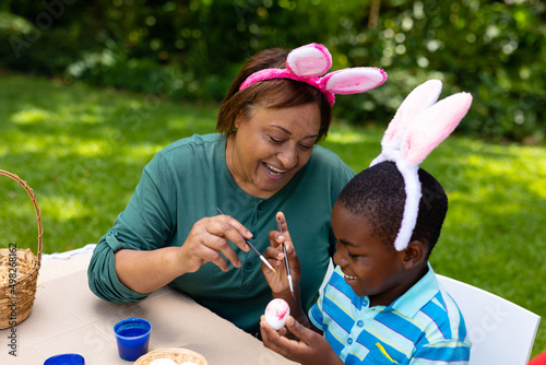 Smiling african american boy and grandmother in bunny ears painting egg at backyard on easter day