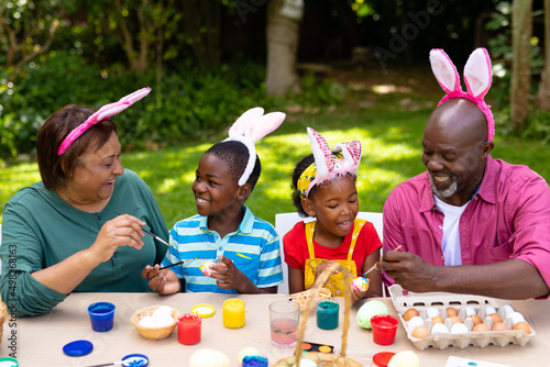 Happy african american siblings and grandparents in bunny ears painting easter eggs in backyard