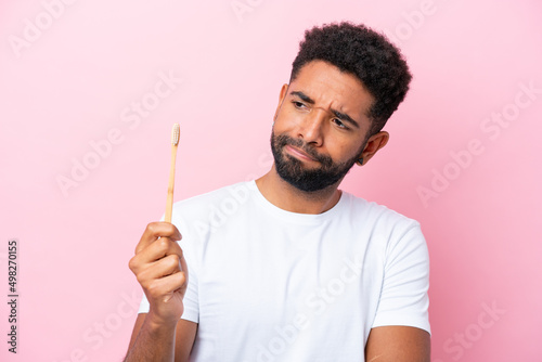 Young Brazilian man brushing teeth isolated on pink background with sad expression