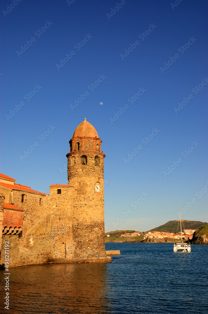 The Church of Our Lady of the Angels in the port of Collioure, France. Moon in the sky and white sailing boat. Sunset.