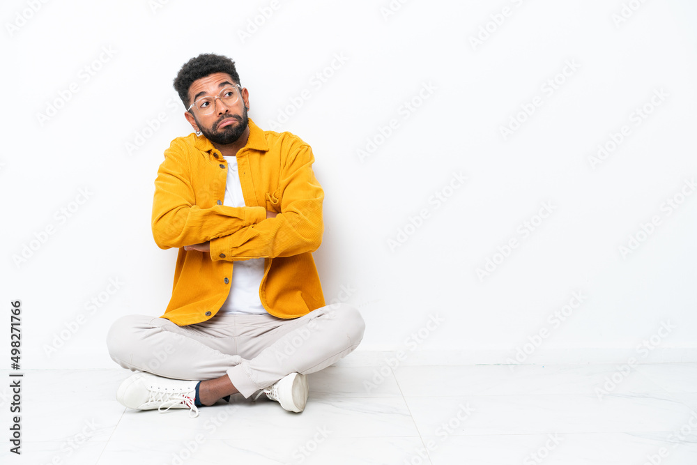 Young Brazilian man sitting on the floor isolated on white background making doubts gesture while lifting the shoulders