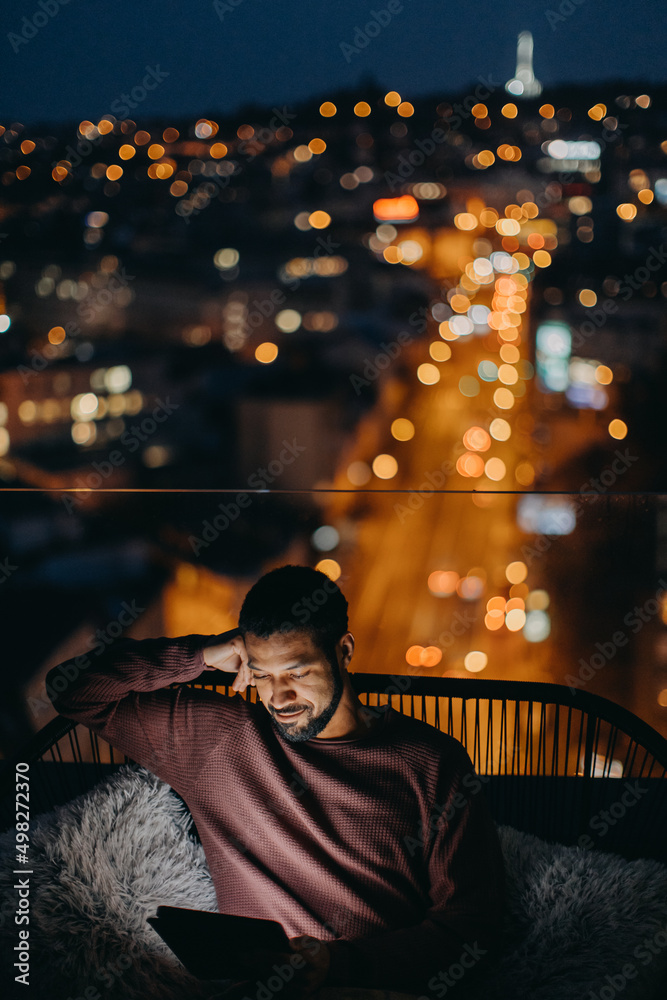Young African American man sitting on balcony with urban view and using tablet at night