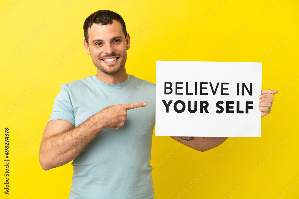 Brazilian man over isolated purple background holding a placard with text Believe In Your Self and pointing it Brazilian man over isolated purple background holding a placard with text Believe In Your Self and pointing it