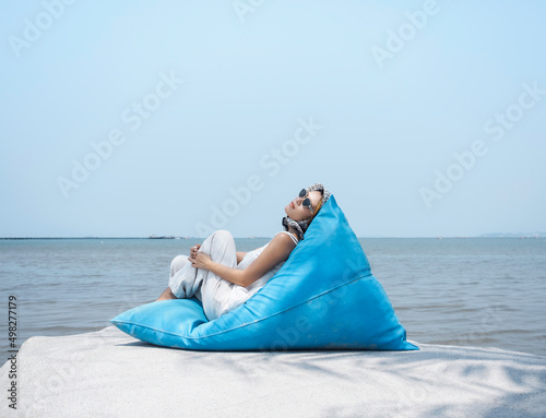 Beautiful Asian woman in casual white shirt and trousers wearing sunglasses and hair scarf reclining on blue bean bag seat on the big rock with sea view and blue sky background on sunny day in summer.