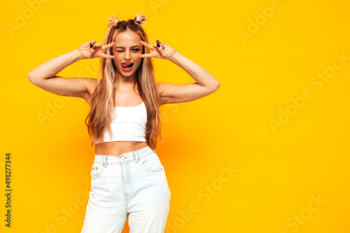 Portrait of young beautiful smiling blond female in trendy summer clothes. Sexy carefree woman posing near yellow wall in studio. Positive model having fun indoors. Cheerful and happy.Shows peace sign