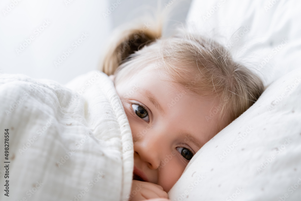 close up view of little girl lying in bed under blanket and looking at camera.