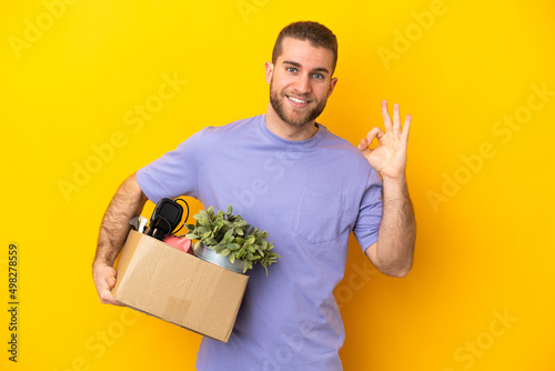 Young caucasian making a move while picking up a box full of things isolated on yellow background showing ok sign with fingers