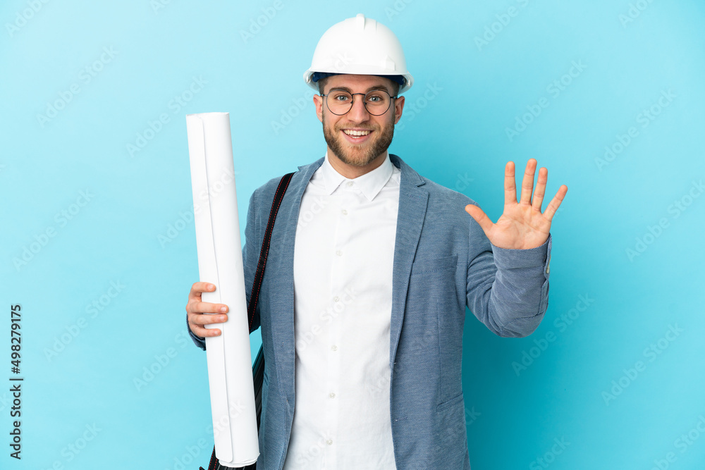 Young architect man with helmet and holding blueprints over isolated background saluting with hand with happy expression