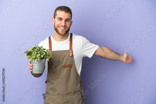 Gardener caucasian man holding a plant isolated on yellow background extending hands to the side for inviting to come