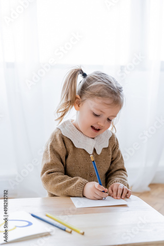joyful child drawing at home near card with blue and yellow heart on table.