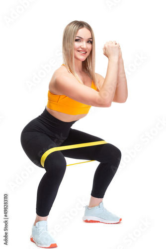 A beautiful, athletic, slim, smiling and cheerful woman in an orange top and black sweatpants performs squats with a fitness band. Lifestyle concept with sports and gym, healthy lifestyle. Isolated on