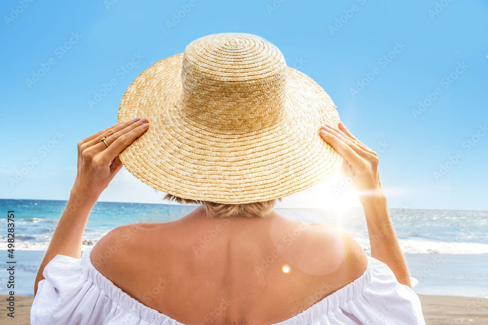 Back view of  a women with straw hat enjoying sunny time at the beach. Tourist girl enjoying breeze at seaside. Travel concept.