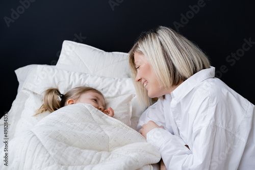 blonde woman smiling near little daughter lying under white blanket.