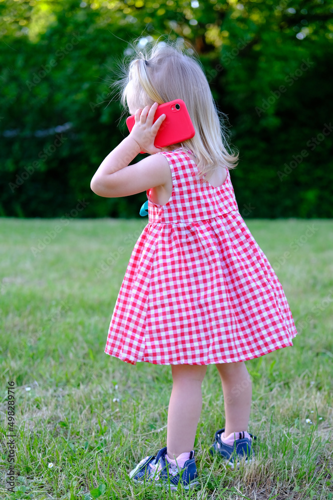 small child, 2-year-old girl with white hair in red dress holds smartphone near her ear, communicates with friends, parents, concept of childhood, previously ability to handle modern technologies small child, 2-year-old girl with white hair in red dress holds smartphone near her ear, communicates with friends, parents, concept of childhood, previously ability to handle modern technologies