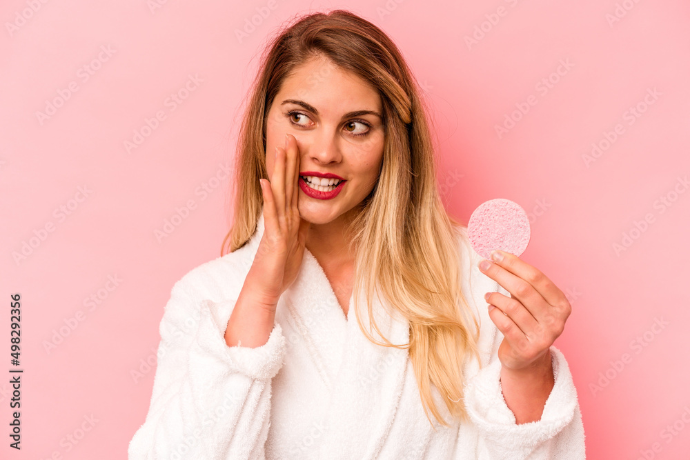 Young caucasian woman holding facial sponge isolated on pink background is saying a secret hot braking news and looking aside Young caucasian woman holding facial sponge isolated on pink background is saying a secret hot braking news and looking aside