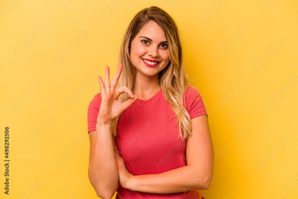 Young caucasian woman isolated on yellow background winks an eye and holds an okay gesture with hand. Young caucasian woman isolated on yellow background winks an eye and holds an okay gesture with hand.