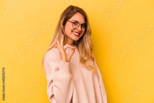 Young caucasian woman isolated on yellow background winks an eye and holds an okay gesture with hand.