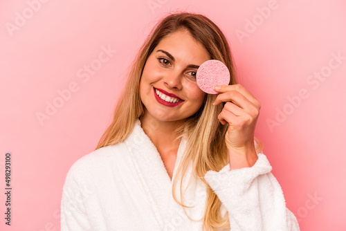 Young caucasian woman holding facial sponge isolated on pink background