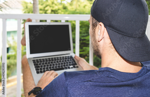 Young man freelancer in cap sitting on balcony with crossed feet and using his laptop, male working on laptop, rear view.