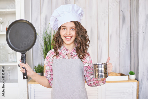 Young girl cooking at kitchen with pan. Curly pretty child portrait. Chef christmas student. Attractive brunette. Children emotion. People baking in cook apron. Close recipe menu