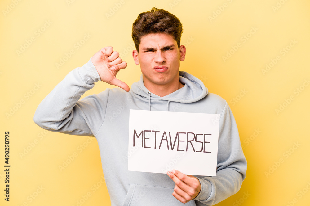 Young caucasian man holding metaverse placard isolated on yellow background showing a dislike gesture, thumbs down. Disagreement concept. Young caucasian man holding metaverse placard isolated on yellow background showing a dislike gesture, thumbs down. Disagreement concept.
