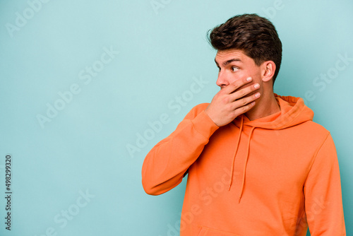 Young caucasian man isolated on blue background thoughtful looking to a copy space covering mouth with hand.