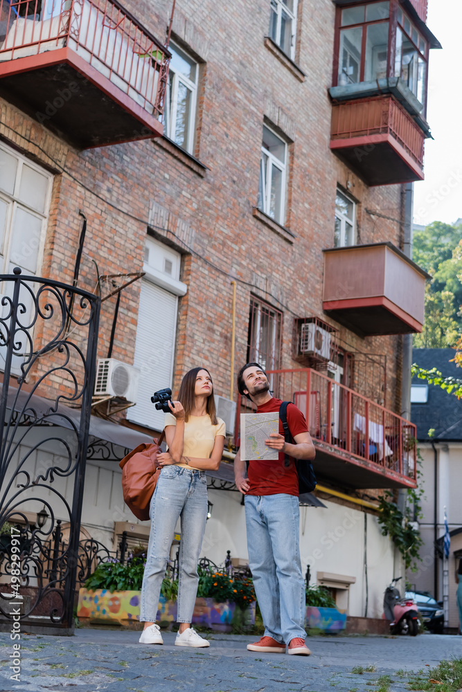 Young couple with map and binoculars looking away near building on urban street.