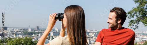 Young woman looking through binoculars near positive boyfriend in viewpoint outdoors, banner.