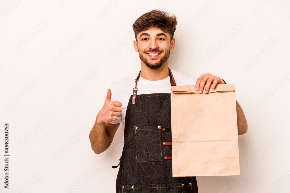 Young hispanic clerk man holding a take away bag isolated on white background smiling and raising thumb up Young hispanic clerk man holding a take away bag isolated on white background smiling and raising thumb up