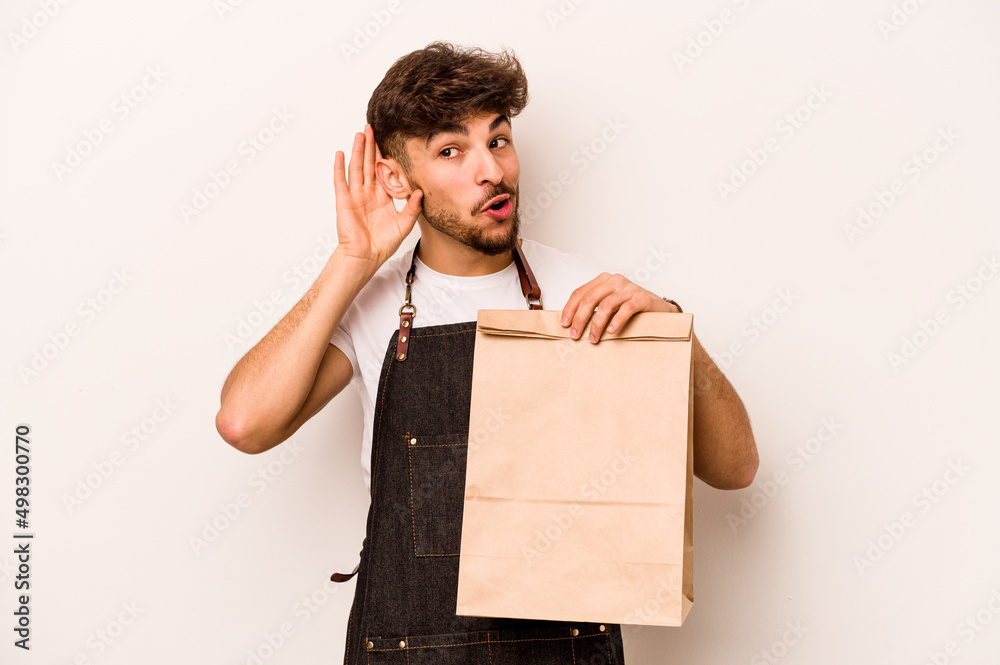 Young hispanic clerk man holding a take away bag isolated on white background trying to listening a gossip. Young hispanic clerk man holding a take away bag isolated on white background trying to listening a gossip.