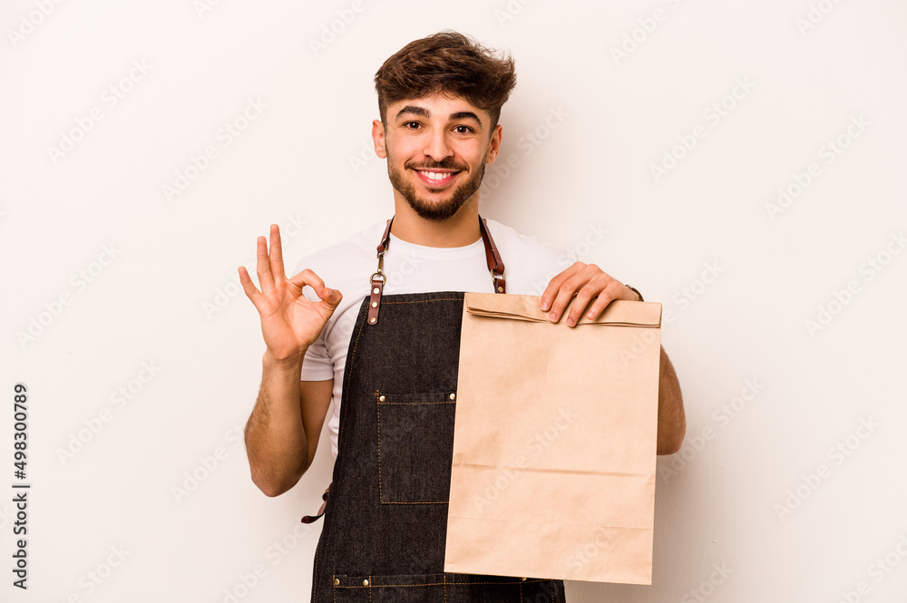 Young hispanic clerk man holding a take away bag isolated on white background cheerful and confident showing ok gesture. Young hispanic clerk man holding a take away bag isolated on white background cheerful and confident showing ok gesture.