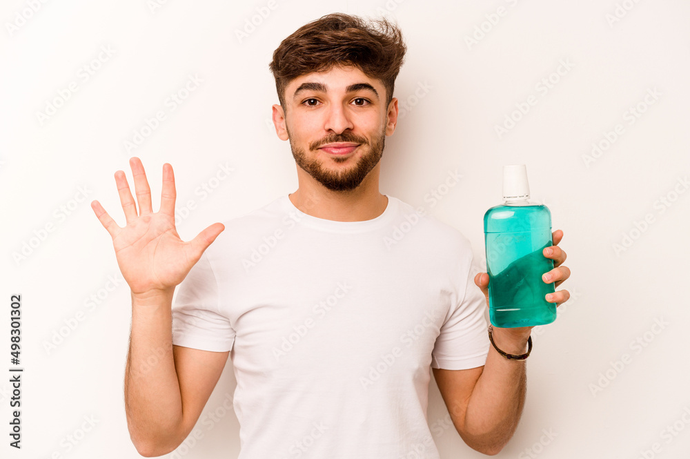 Young hispanic man holding mouthwash isolated on white background smiling cheerful showing number five with fingers. Young hispanic man holding mouthwash isolated on white background smiling cheerful showing number five with fingers.