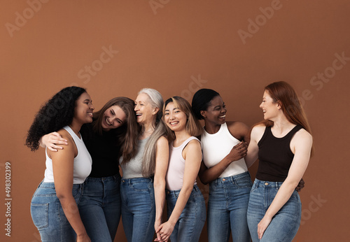 Six laughing women of different body types and ethnicities in studio. Group portrait of smiling females standing together against brown background.