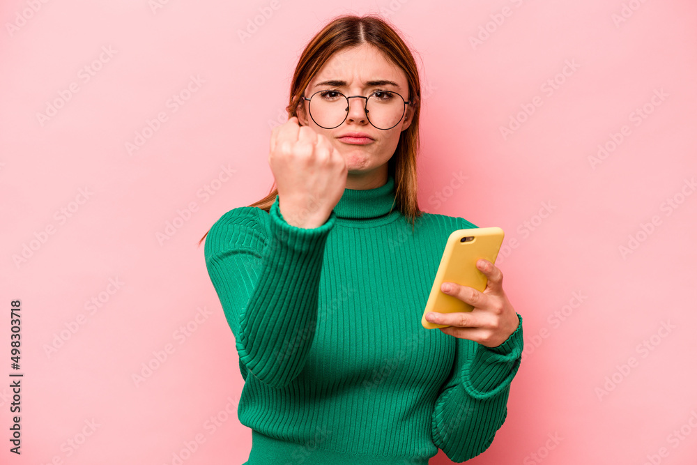 Young caucasian woman holding mobile phone isolated on pink background showing fist to camera, aggressive facial expression.