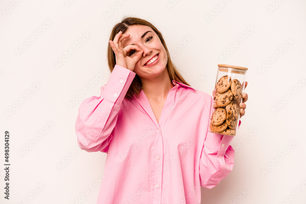 Young caucasian woman holding cookies jar isolated on white background excited keeping ok gesture on eye. Young caucasian woman holding cookies jar isolated on white background excited keeping ok gesture on eye.
