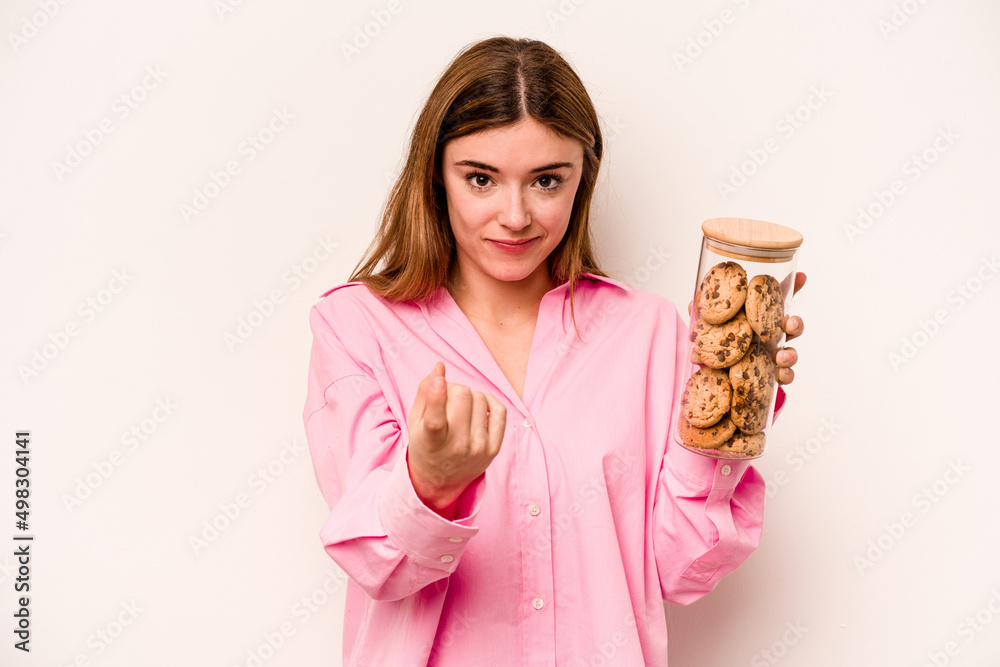 Young caucasian woman holding cookies jar isolated on white background pointing with finger at you as if inviting come closer. Young caucasian woman holding cookies jar isolated on white background pointing with finger at you as if inviting come closer.