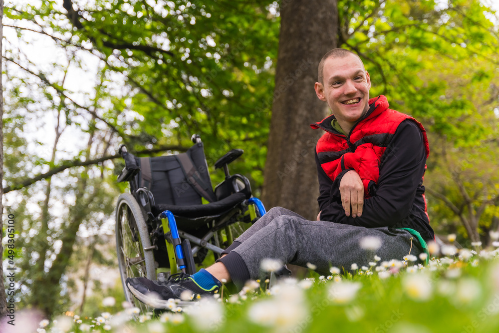 Fototapeta premium Portrait of a person with a disability sitting on the grass next to the wheelchair in a daisy flower, smiling and enjoying nature