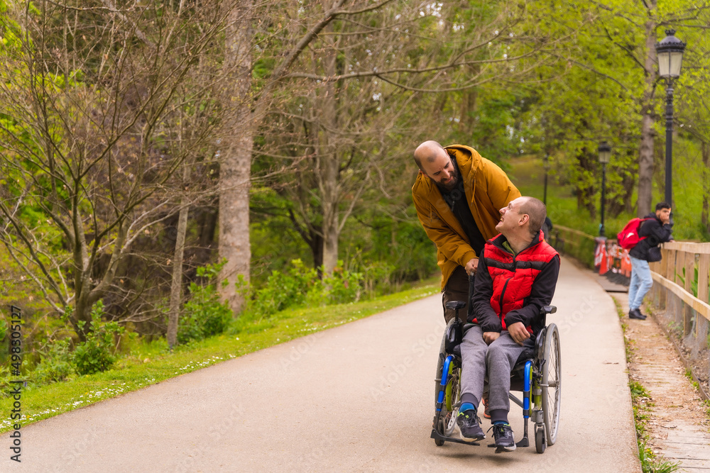 A person with a disability in a wheelchair being pushed by a friend in a public city park, strolling along a path A person with a disability in a wheelchair being pushed by a friend in a public city park, strolling along a path