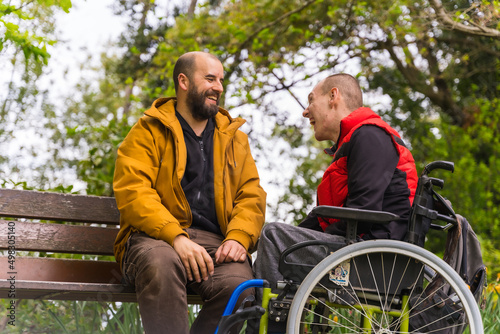 A person with a disability young man in a wheelchair with a friend on a bench in a public park in the city, talking and laughing photo