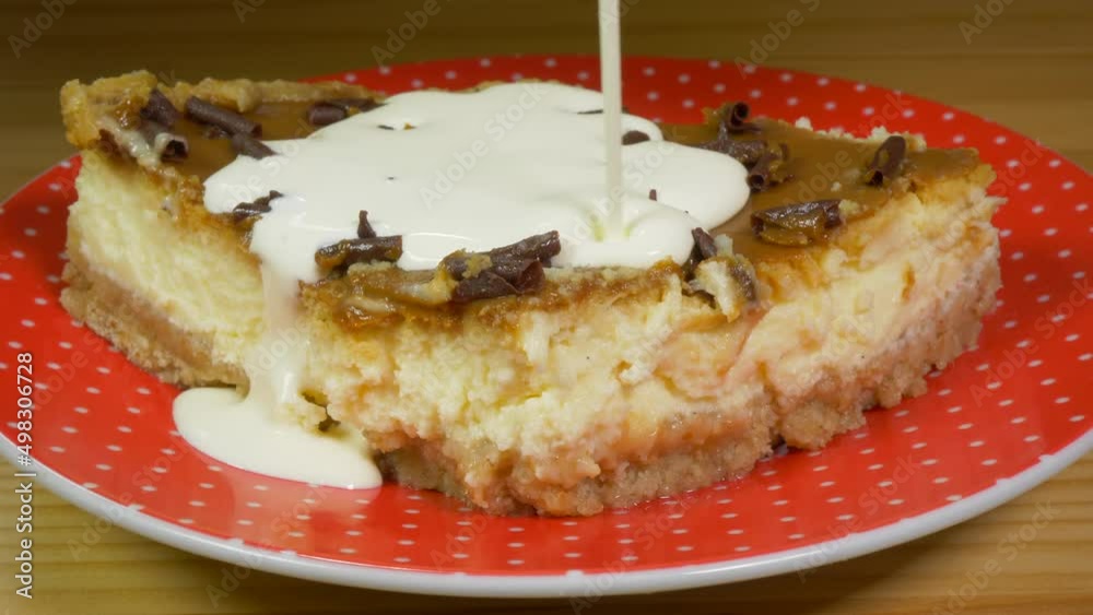 Closeup of dairy cream being poured over a slice of cheesecake, topped with a layer of caramel toffee and finished with rolled chocolate pieces, on a red plate. Closeup of dairy cream being poured over a slice of cheesecake, topped with a layer of caramel toffee and finished with rolled chocolate pieces, on a red plate.