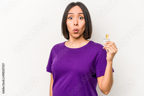 Young hispanic pregnant woman holding pacifier isolated on white background shrugs shoulders and open eyes confused.