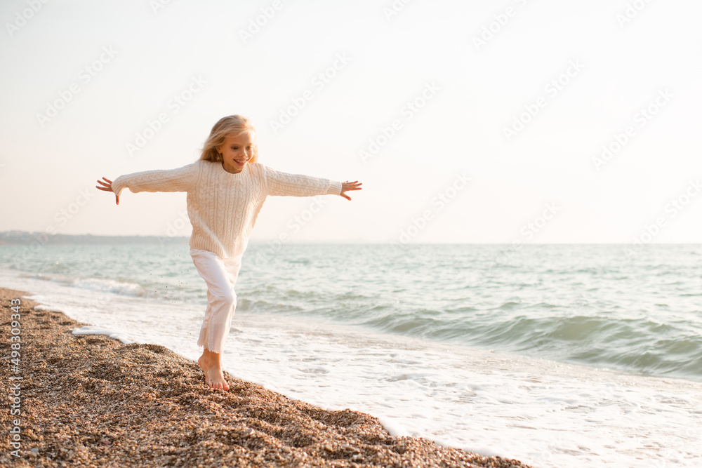 Happy smiling kid girl 5-6 year old wear knit sweater and pants running over sea shore outdoors. Cheerful little toddler having fun at beach. Summer season. Childhood. Happiness. Happy smiling kid girl 5-6 year old wear knit sweater and pants running over sea shore outdoors. Cheerful little toddler having fun at beach. Summer season. Childhood. Happiness.