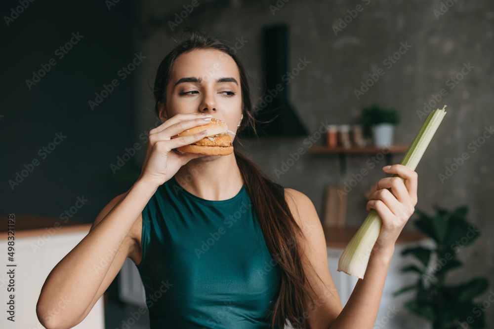 Fototapeta premium Hungry woman eating burger, holding vegetable, standing in the kitchen. Choosing between healthy and unhealthy food. Sporty young girl. Holding hands. Fast food