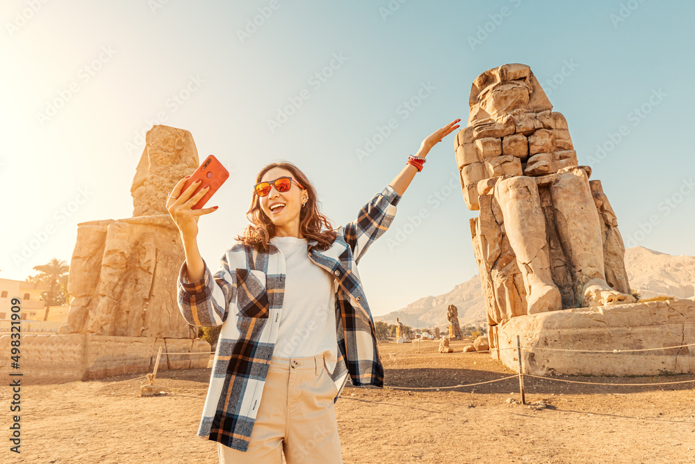 Travel blogger girl takes selfie pictures on a smartphone at the famous two Colossi of Memnon - massive ruined statues of the Pharaoh Amenhotep III. World tourism attractions