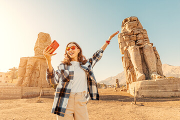 Travel blogger girl takes selfie pictures on a smartphone at the famous two Colossi of Memnon - massive ruined statues of the Pharaoh Amenhotep III. World tourism attractions